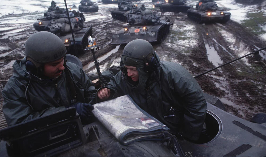 Two men reading booklet on top of military car surrounded by tank cars in mud.
