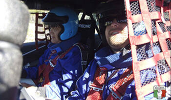 Two men inside racing car with racing gear and helmet.