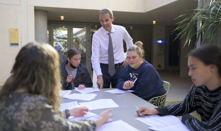 Candid photo of Daniel Krauss standing at an outdoor class setting with students seated around a table with papers.