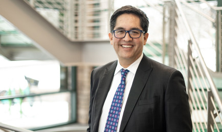 Portrait of Alex Hernandez ’96 in a black suit, standing by a sunlit stairway.